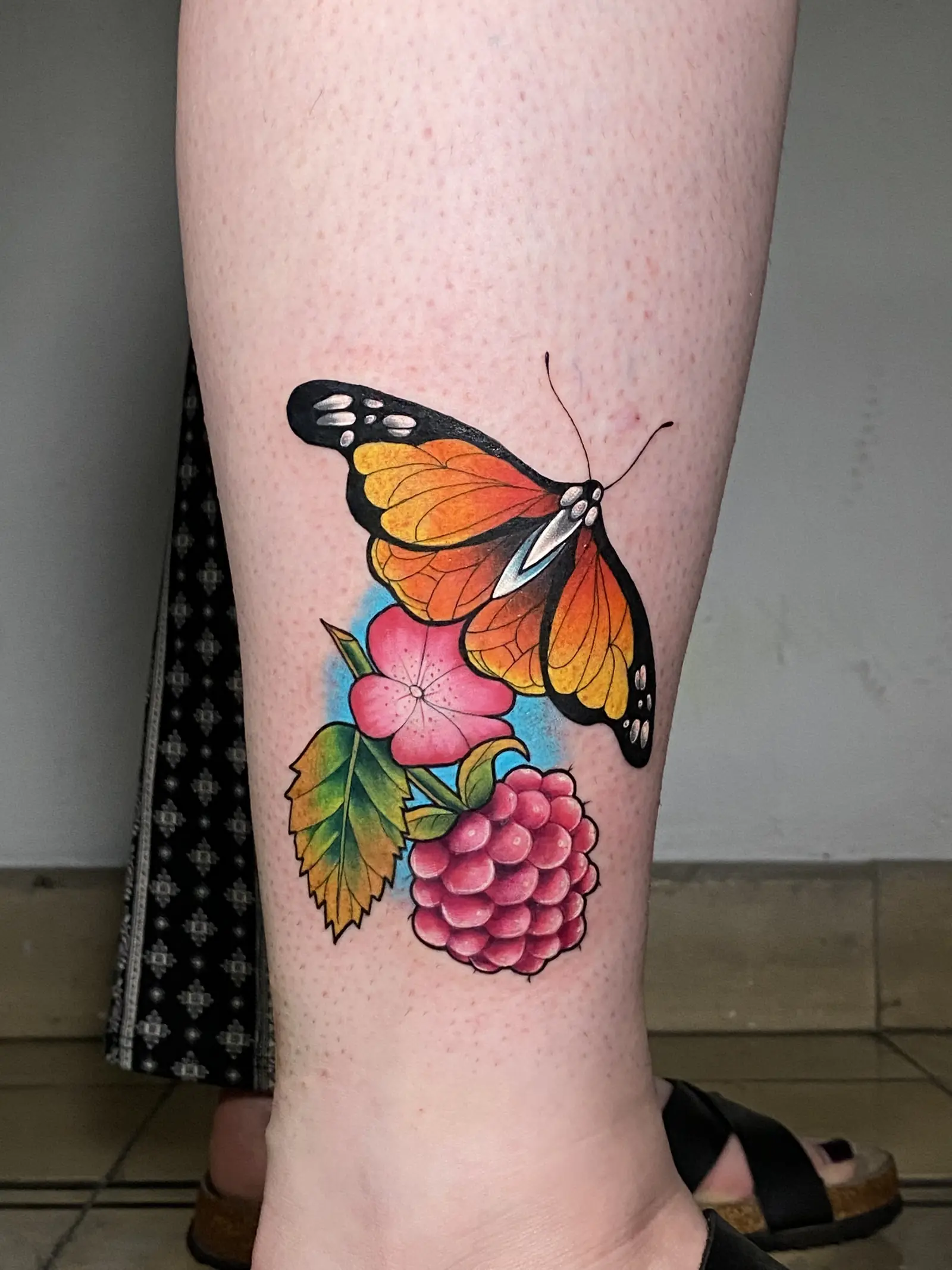 Tatuaje a color de una mariposa, flores y frambuesas en la pantorrilla, hecho en Cartagena