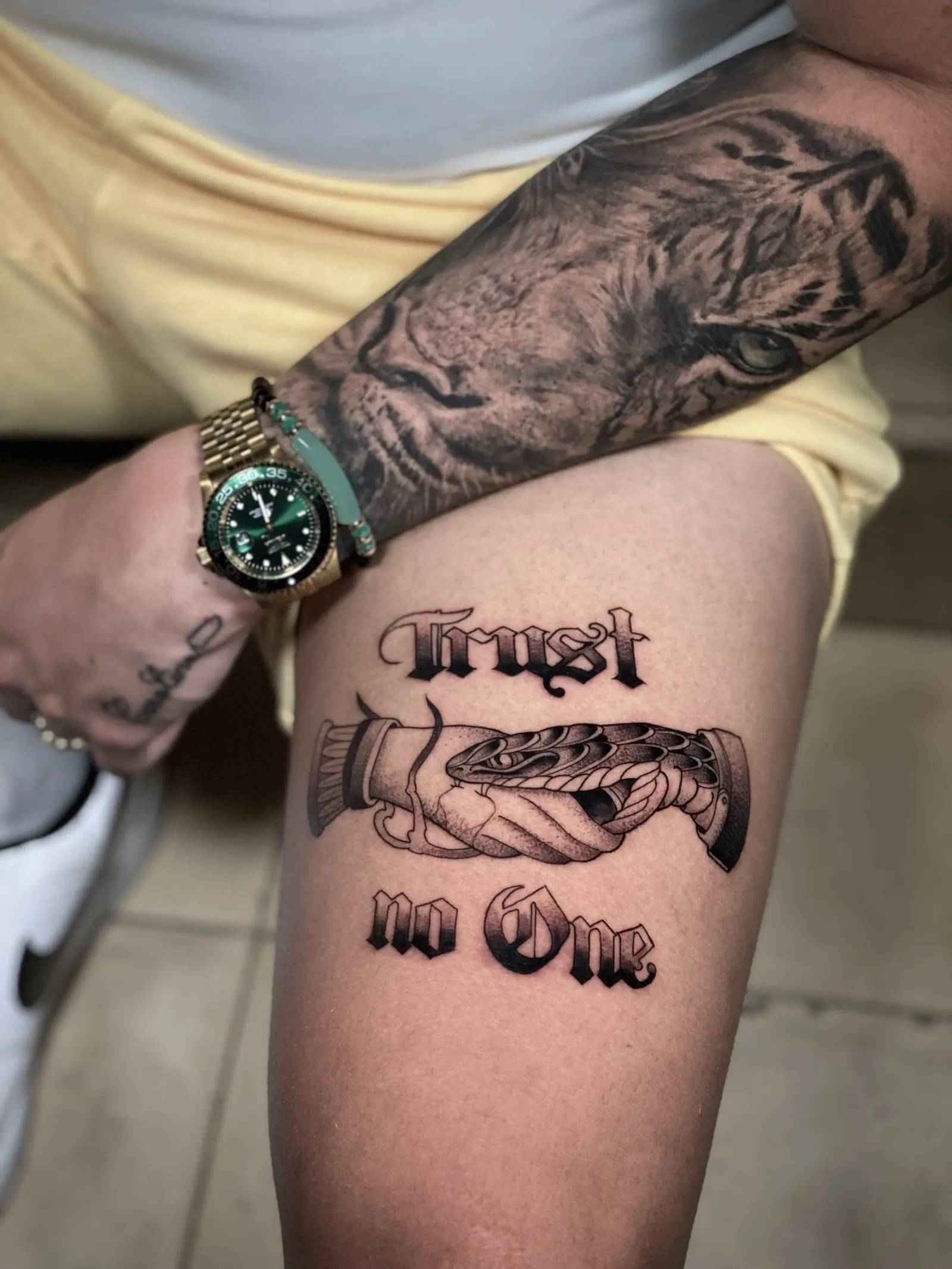 Tatuaje con la frase "Trust no one" en letra gótica, con una serpiente y un apretón de manos en el muslo, hecho en Cartagena